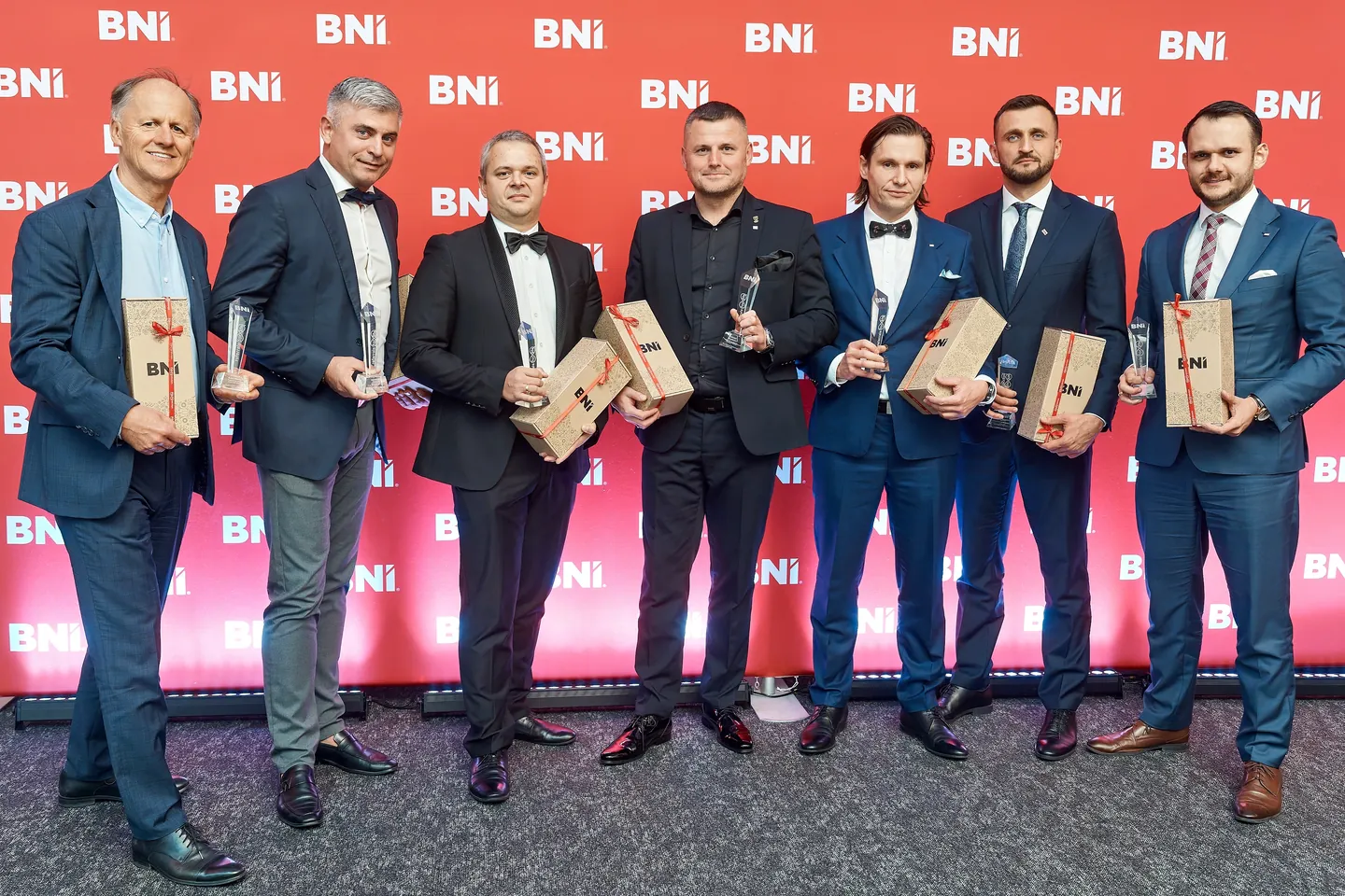 Siedmiu laureatów BNI Awards z nagrodami i upominkami, na tle ścianki z logotypami BNI.