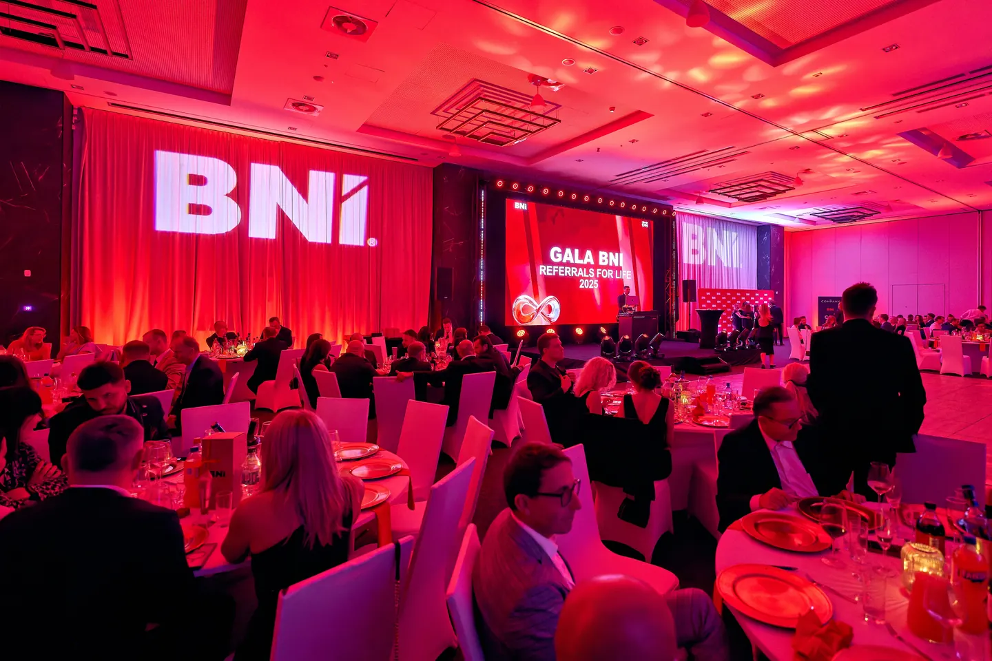 Sala gali BNI Referrals for Life w czerwonym świetle: Goście przy okrągłych stołach, projekcje logotypu BNI i ekran sceniczny.