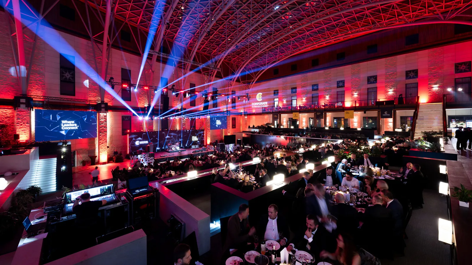 Wieczorna gala Business Weekend Corporate Connections: setki gości przy stołach w hali z kratownicowym sklepieniem, czerwono-niebieskie światła sceniczne i ekran z hasłem Where Leaders Connect.