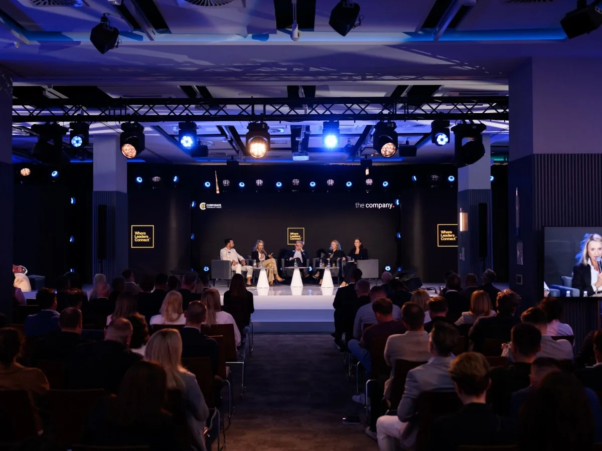 Panel na scenie Business Weekend: pięcioro rozmówców w fotelach, pełna widownia i niebieskie światła sceniczne, w tle logotypy Corporate Connections i the company.