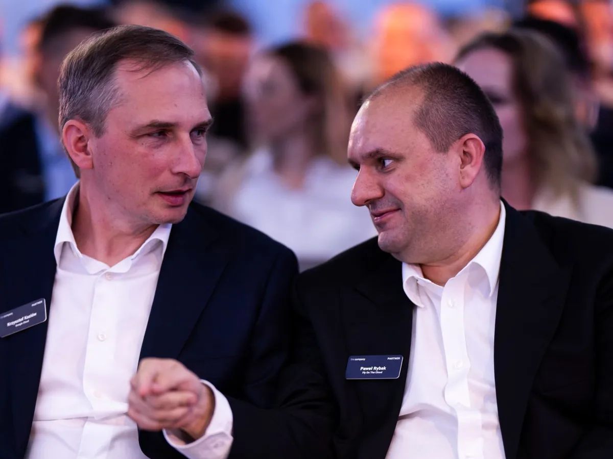 Dwaj Przedsiębiorcy w garniturach rozmawiają półgłosem na widowni, w tle rozmyta sala.