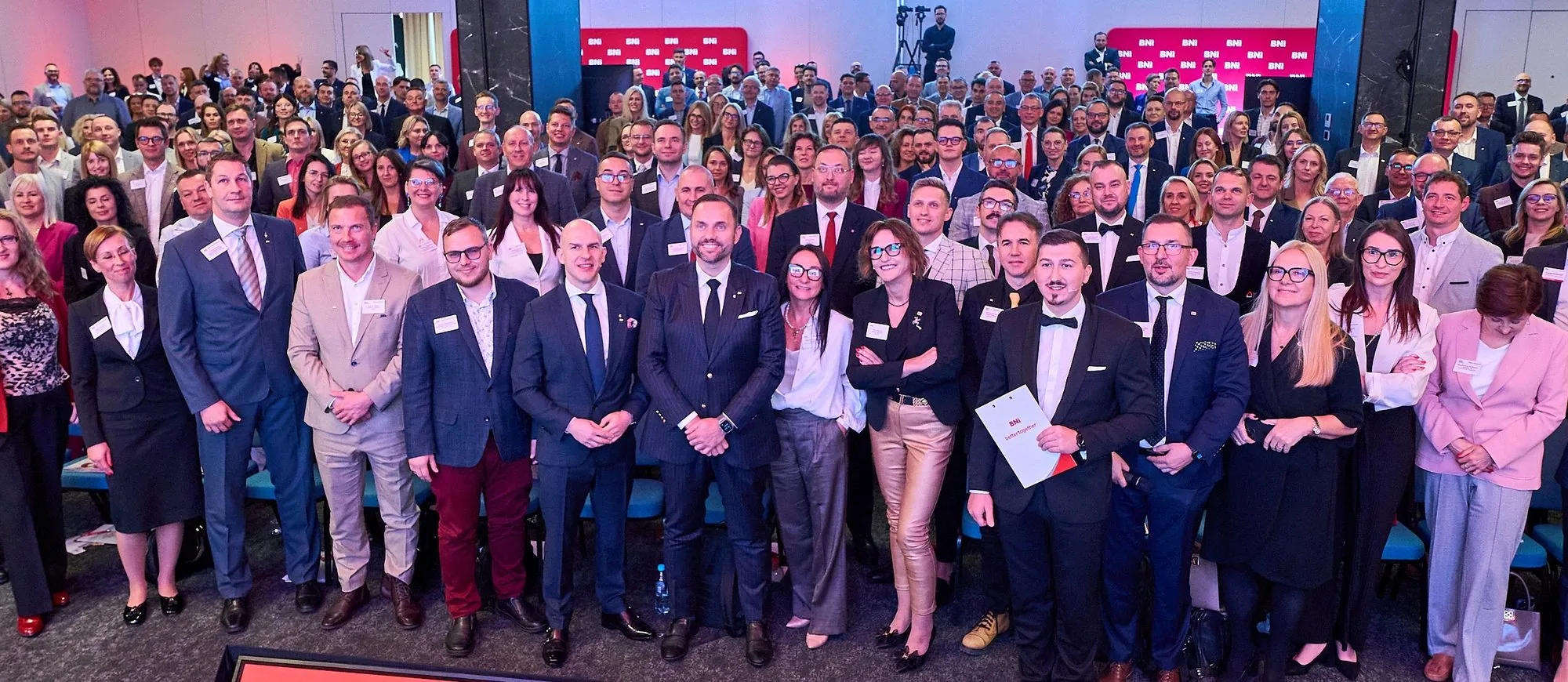 Kilkuset przedsiębiorczyń i przedsiębiorców z BNI pozuje razem w pełnej sali konferencyjnej, w tle ścianka z logotypami BNI.