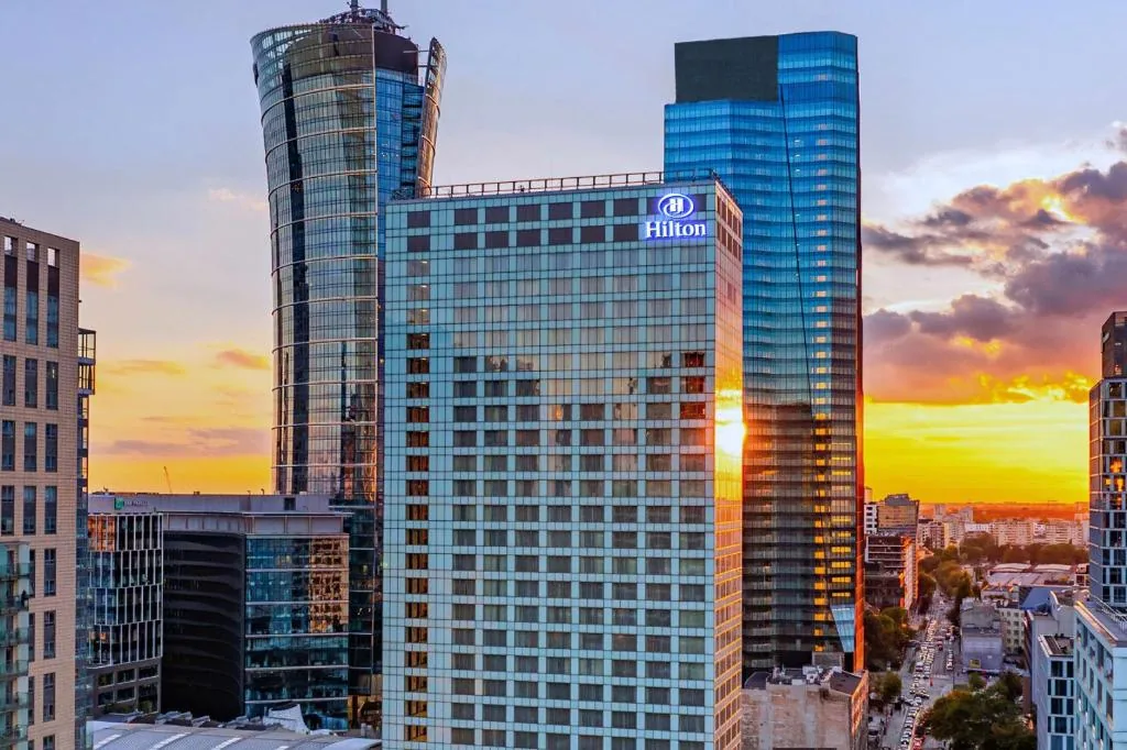 Hotel Hilton Warsaw City z podświetlonym logo, na tle wieżowców i zachodu słońca.