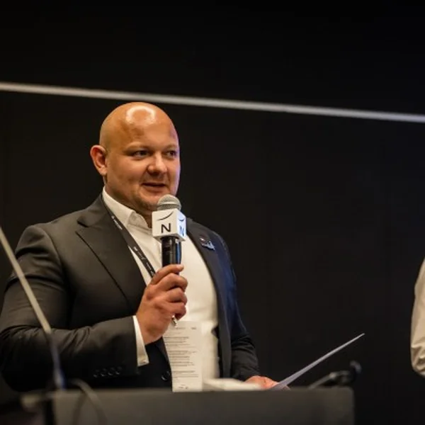 Paweł Pawul z mikrofonem i kartkami w dłoni, w ciemnej marynarce, podczas wystąpienia.