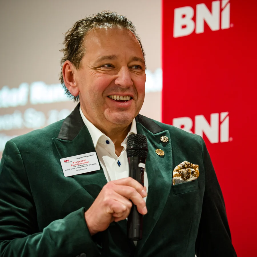 Krzysztof Rumianowski z mikrofonem podczas wystąpienia, w zielonej aksamitnej marynarce, w tle ścianka z logotypami BNI.