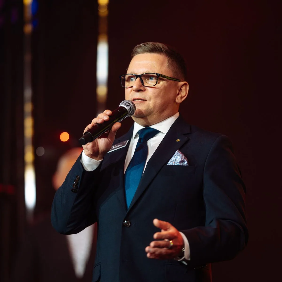 Maciej Tymusz w granatowym garniturze na scenie, z mikrofonem.